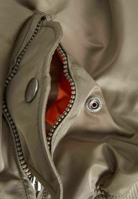 Poche de veste beige partiellement ouverte avec fermeture éclair métallique visible, bouton-pression et doublure intérieure orange vif.