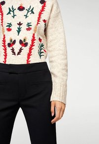 Maglione beige lavorato a maglia con ricami floreali colorati in rosso, verde e blu navy, abbinato a pantaloni neri su misura con tasche laterali.