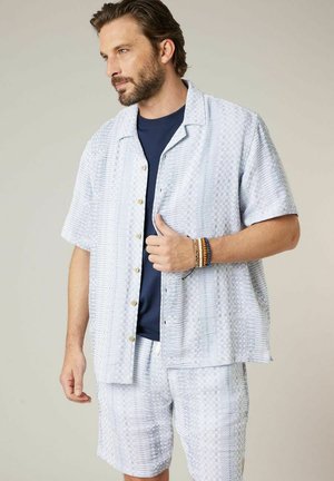 Homme portant une chemise légère à manches courtes à motifs et un short assorti, un T-shirt bleu marine en dessous, et des bracelets perlés au poignet gauche.