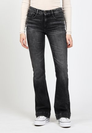 Woman wearing dark gray bootcut jeans, white long-sleeve top, and white sneakers, standing against a plain background.