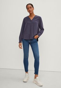 Blusa lunga viola con scollo a V e vestibilità morbida, abbinata a jeans blu attillati e sneakers bianche, su uno sfondo neutro.