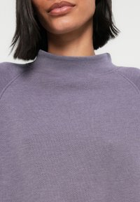 Purple knitted sweater with a high collar, featuring a soft texture and raglan sleeves. Close-up view focuses on the neckline and fabric details.