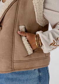 Bruine fleece-voering vest met sherpa kraag en zijzakken. Beige textuur lange mouwen shirt eronder. Gouden armbanden om de pols.