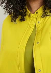 Světlá žlutá bunda s tlakovým uzávěrem a vysokým límcem, která má hladký povrch a vrstvený zelený interiér.