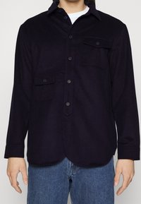 Navy button-up shirt with long sleeves, two front pockets, and a rounded hem. Made of a soft, textured fabric. Subtle branding on left pocket.