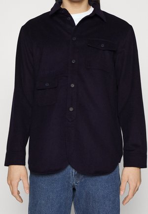 Camisa de botones azul marino con mangas largas, dos bolsillos frontales y dobladillo redondeado. Confeccionada en una tela suave y texturizada. Marca sutil en el bolsillo izquierdo.
