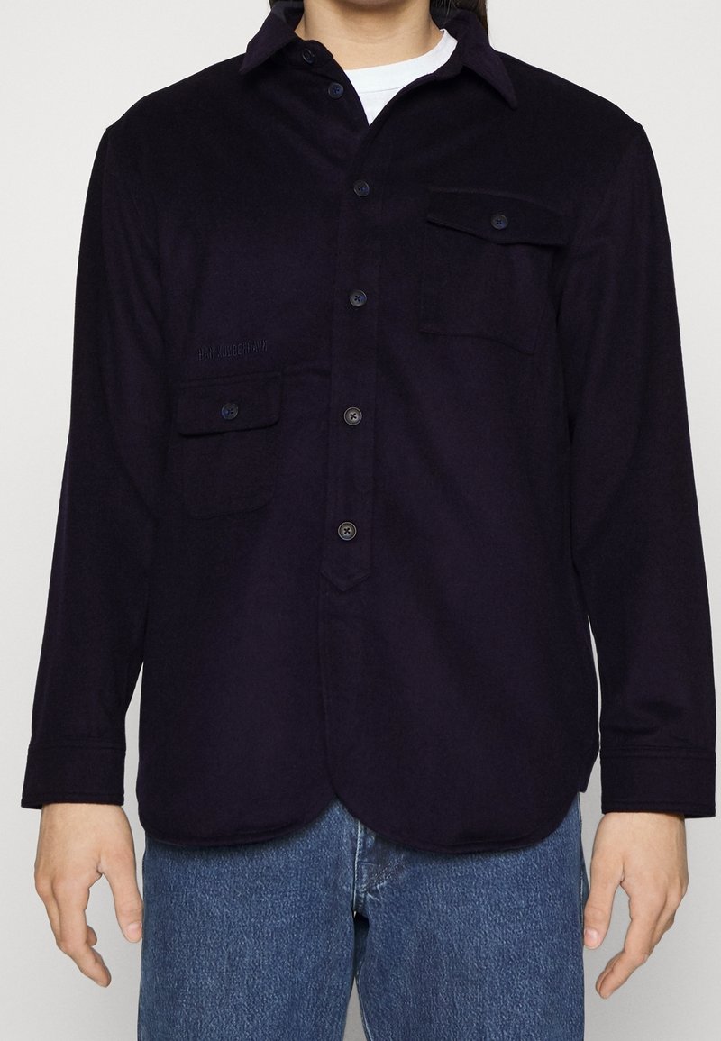 Navy button-up shirt with long sleeves, two front pockets, and a rounded hem. Made of a soft, textured fabric. Subtle branding on left pocket.