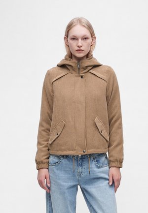 Frau trägt eine beige Kordjacke mit Kapuze, Reißverschluss und Knopfverschluss sowie zwei vorderen Taschen mit Knopfklappen, kombiniert mit hellblauen Jeans.