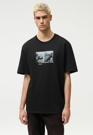 Młody mężczyzna z krótkimi blond włosami nosi czarny, oversize'owy t-shirt z rozmytym czarno-białym nadrukiem i napisem "DIVERSE SYSTEM".