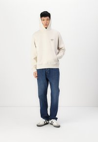 Lichtbeige hoodie met een voorzak en een klein logo. Gecombineerd met blauwe jeans en witte sneakers met zwarte accenten.