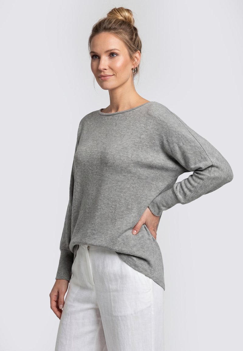 Femme aux cheveux blonds attachés en chignon, portant un pull gris à manches longues et un pantalon blanc, debout avec une main sur la hanche sur un fond uni.