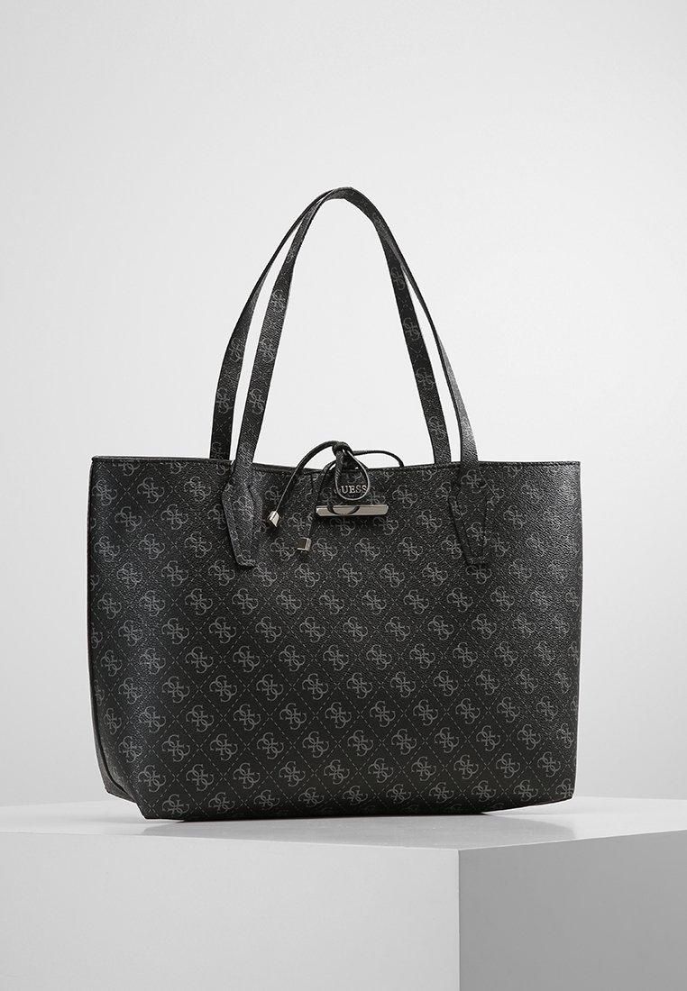 Sac cabas noir avec un motif monogramme sur l'ensemble, double poignée d'épaule, et une petite étiquette circulaire de la marque Guess attachée avec un lacet en cuir.