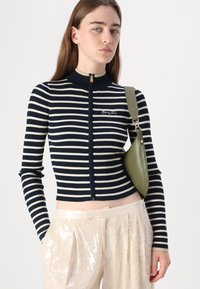 Navy and cream striped ribbed sweater with a high collar and zip closure, paired with beige sequined pants and a green handbag.