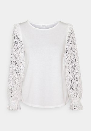 Haut blanc à manches longues avec col rond et manches transparentes en dentelle florale, doté de poignets à volants et de légers détails froncés aux épaules.