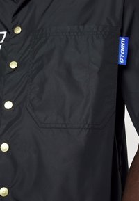 Veste noire avec une grande poche avant et des boutons-pression dorés, avec une étiquette en tissu bleu portant le mot « STORM » sur le côté.