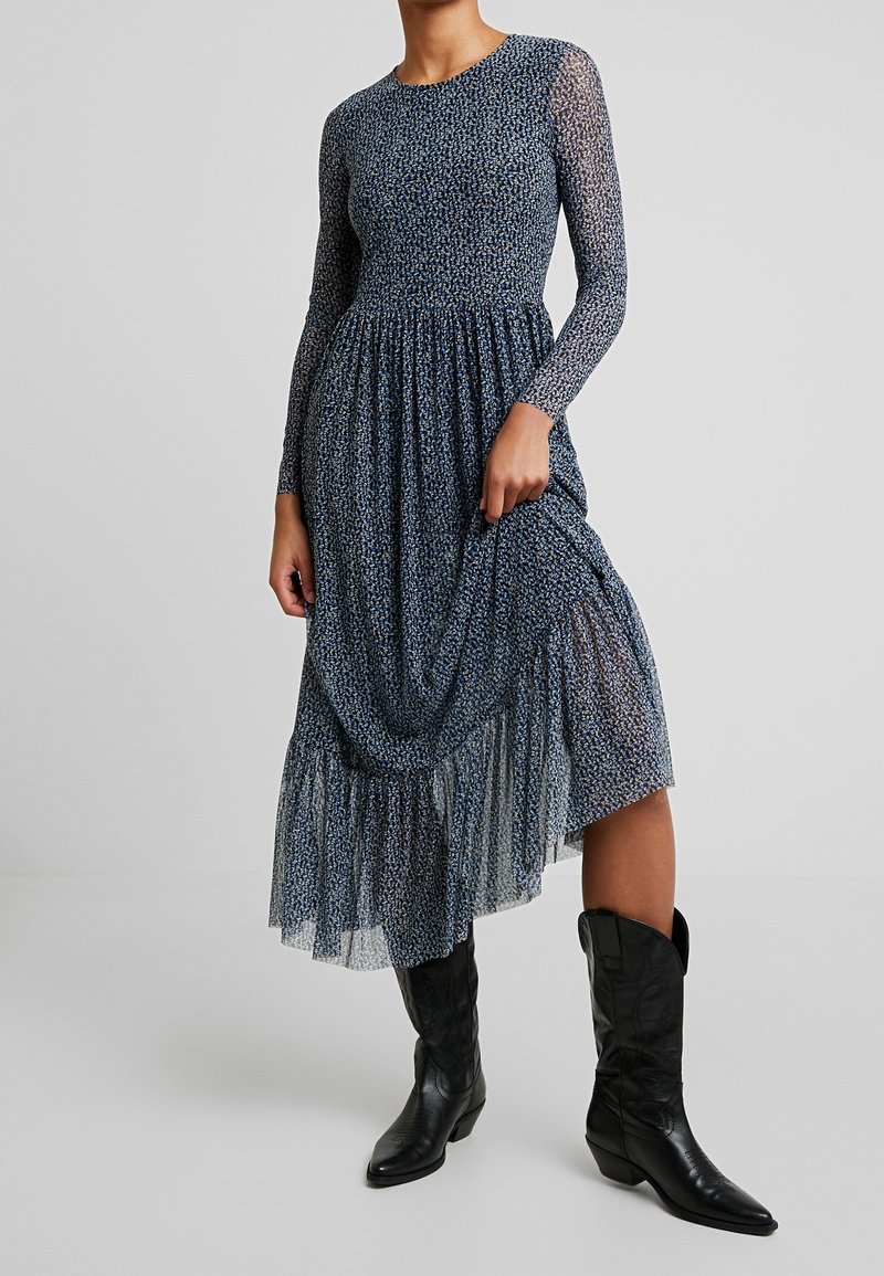 Femme portant une robe midi bleue fleurie à manches longues transparentes et des bottes en cuir noires mi-mollet, tenant une partie de la robe.