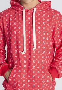 Sudadera roja con capucha que presenta un patrón repetido de logotipos blancos, bolsillo canguro en la parte delantera y cordones. Los puños y el dobladillo acanalados completan el diseño.