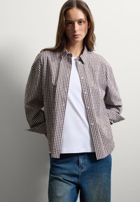 Mujer con el cabello largo que lleva una camisa de cuadros marrón y blanca sobre una camiseta blanca, jeans azules, de pie con las manos detrás de la espalda.