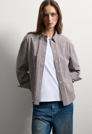 Femme aux longs cheveux portant une chemise à carreaux marron et blanc sur un t-shirt blanc, un jean bleu, se tenant les mains derrière le dos.