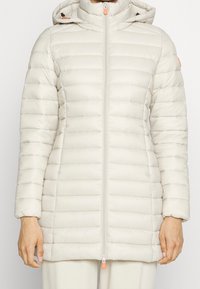 Lys beige pufferjakke med horisontalt quiltet design, opretstående krave, lynlås og kontrastfarvede orange hardware-detaljer.