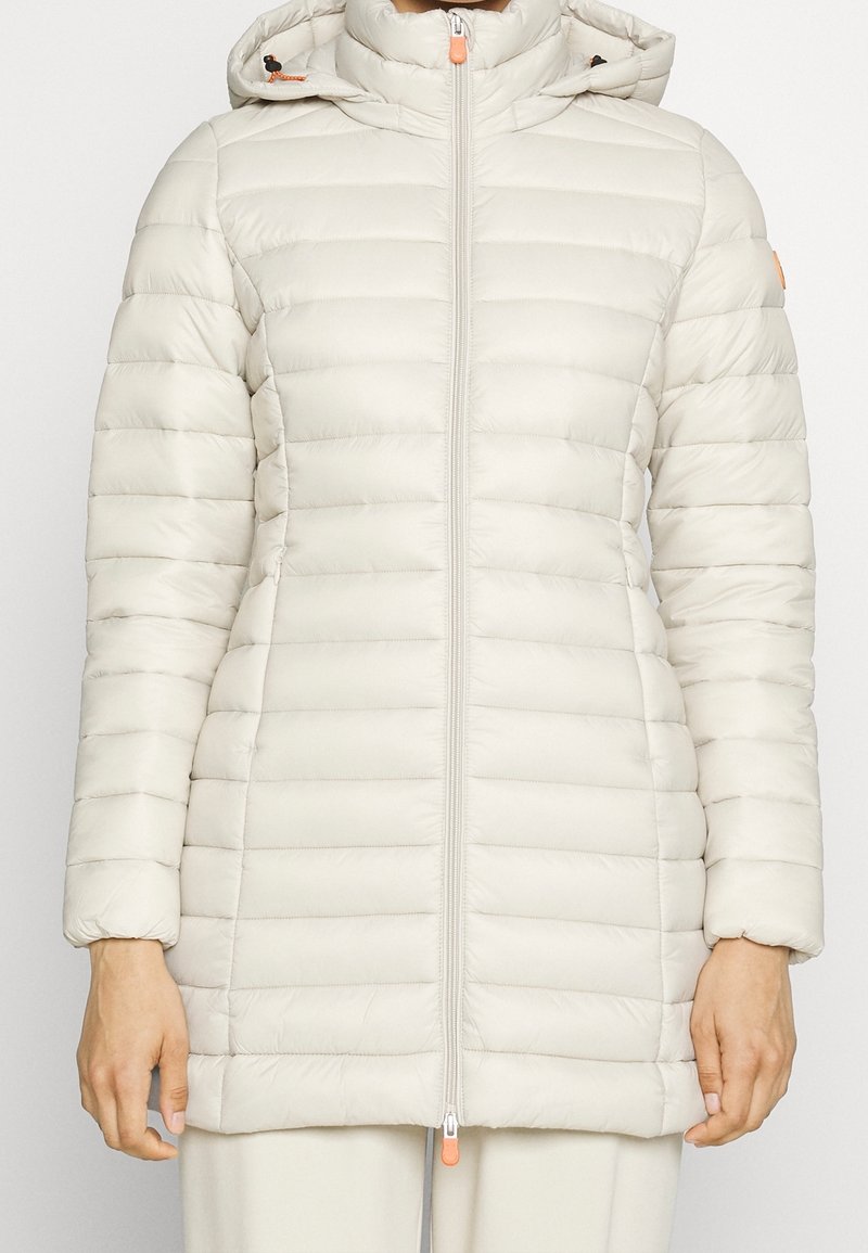 Lys beige pufferjakke med horisontalt quiltet design, opretstående krave, lynlås og kontrastfarvede orange hardware-detaljer.