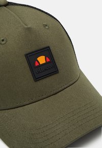 Casquette vert olive en tissu texturé, dotée d'un patch logo rectangulaire noir avec un design rouge et jaune à l'avant.