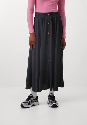 Femme portant une jupe midi noire à boutons sur le devant, un haut rose à manches longues, des chaussettes blanches et des baskets noires et blanches, debout devant un fond uni.