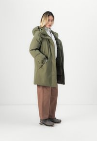 Olive green, knee-length parka with a hood, zip front, and elastic cuffs. Worn over a gray sweater and brown pants, with gray shoes.