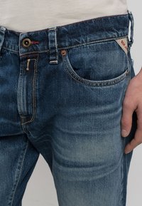 Blå denimjeans med en avslappnad passform, som har flera fickor, knappstängning och subtila blekningar nära låren och knäna.