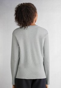 Graues Langarmshirt aus weichem Stoff, mit rundem Halsausschnitt und nahtdetails. Getragen mit schwarzen Hosen, von hinten betrachtet.