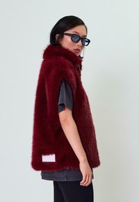 Bordo dirbtinio kailio liemenė su aukštu apykakle, be rankovių dizainu ir baltu etikete apatinėje nugaros dalyje, dėvima ant tamsios marškinėlių ir juodų kelnių.