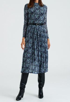 Pitkähihainen, tummansininen mekko, jossa on vihreä- ja valkoispainanteinen paisley-kuvio, vyötäröltä vyötetty mustalla vyöllä, yhdistetty polveen asti ulottuviin mustiin saappaisiin.