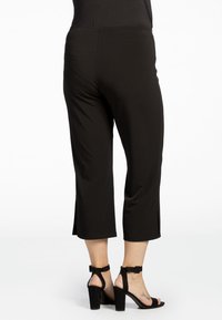 Zwarte cropped broek van rekbaar materiaal, met een gladde textuur en zijsplitten aan de zoom, gecombineerd met zwarte haksandalen.