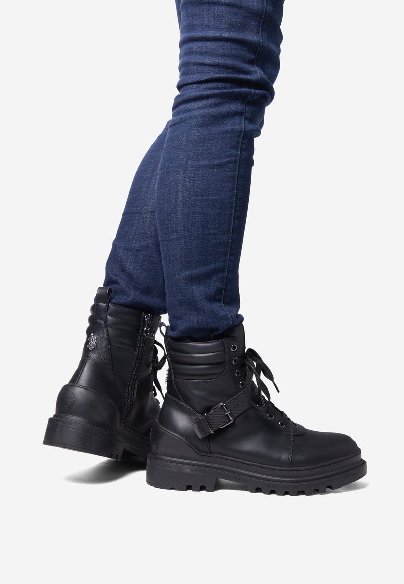 Botas de combate de piel negra con parte frontal con cordones, cuello acolchado, cremallera lateral y correa ajustable. Suela de goma texturizada con patrón de tacos.
