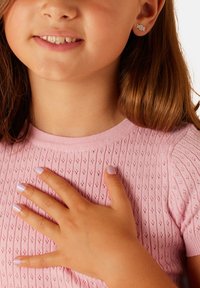 Lichtroze gebreide top met korte mouwen en een gestructureerd patroon. Een hand rust op de borst, met lila geschilderde nagels. Kleine studoorbellen zijn zichtbaar.