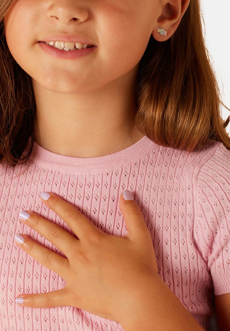 Lichtroze gebreide top met korte mouwen en een gestructureerd patroon. Een hand rust op de borst, met lila geschilderde nagels. Kleine studoorbellen zijn zichtbaar.