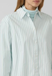 Vrouw in een lichtblauw en wit verticaal gestreept overhemd met knopen, gedragen over een lichtblauwe turtleneck, met lang steil haar zichtbaar.
