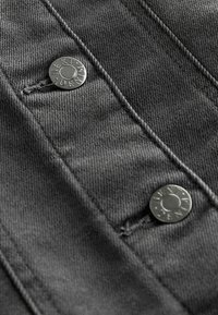 Detailní záběr tmavé džínoviny se dvěma kovovými knoflíky s vyrytým nápisem "NEXT DENIM" a viditelnými švy.