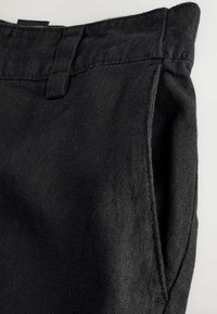 Gros plan sur un pantalon en tissu noir montrant la ceinture, la passant de ceinture, la couture latérale et l'ouverture de la poche sur un fond clair.