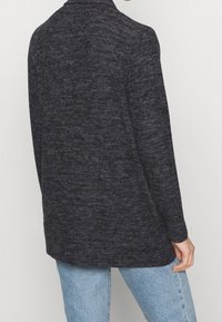 Cardigan in maglia grigio scuro a maniche lunghe con un motivo testurizzato, apertura frontale e orlo posteriore leggermente allungato. Abbinato a jeans azzurri chiari.