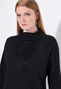 Schwarzes Langarmoberteil mit hohem Ausschnitt, das ein strukturiertes, mit Pailletten besetztes Schleifenmotiv an der linken Schulter aufweist. Weicher Stoff, einfache Silhouette.
