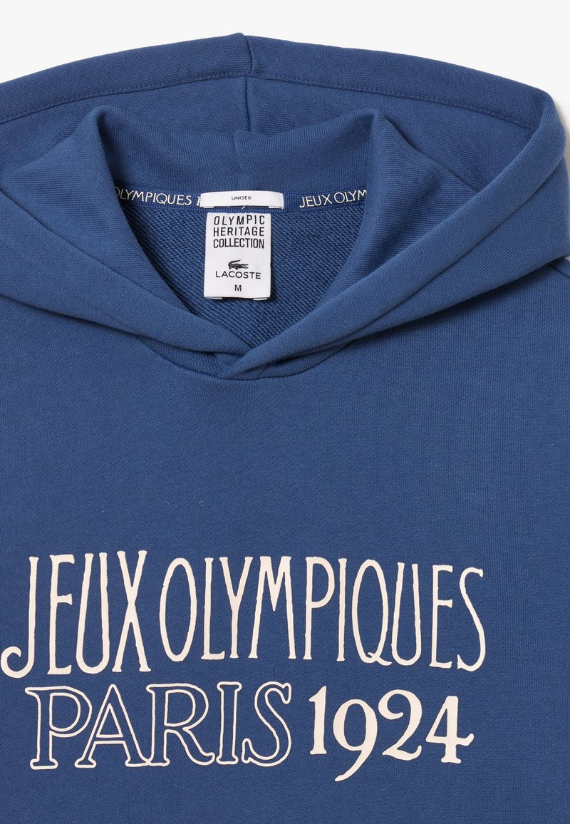 Blaue Kapuzenjacke mit Kängurutasche und weißem Text: "JEUX OLYMPIQUES PARIS 1924." Etikett zeigt "OLYMPIC HERITAGE COLLECTION" und Größe M.