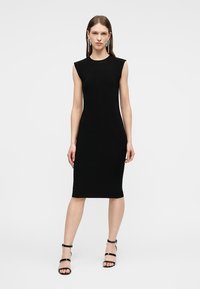 Γυναίκα που φοράει μαύρο φόρεμα χωρίς μανίκια μέχρι το γόνατο, μαύρα πέδιλα με λουράκια και μακριά κρεμαστά σκουλαρίκια, στέκεται μπροστά σε απλό φόντο.