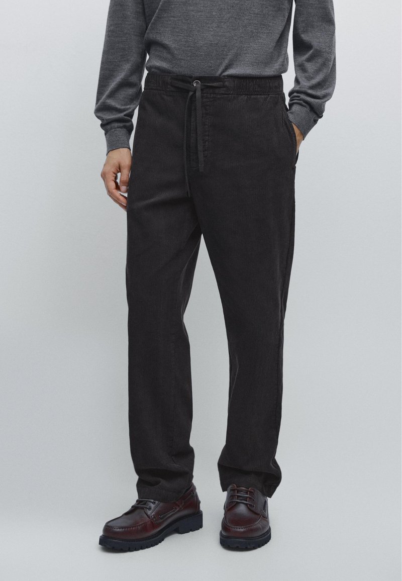 Black corduroy pants with a relaxed fit, elastic waistband, and drawstring; paired with dark leather shoes. Textured fabric detail visible.