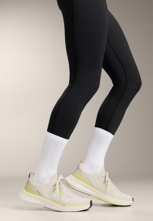 Fekete leggingset viselő lábak, fehér bokazokniban és világos bézs futócipőben, semleges háttér előtt.