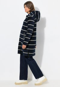 Cappotto con cappuccio a righe blu marina e strisce bianche, dotato di chiusura con zip e tasche laterali. Abbinato a jeans scuri e sneakers bianche chunky.
