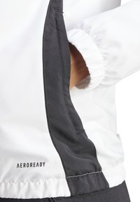 Aeroready-jack in een witte buitenlaag met een contrasterende zwarte zijstreep. Het heeft elastische manchetten en een gladde, matte textuur.