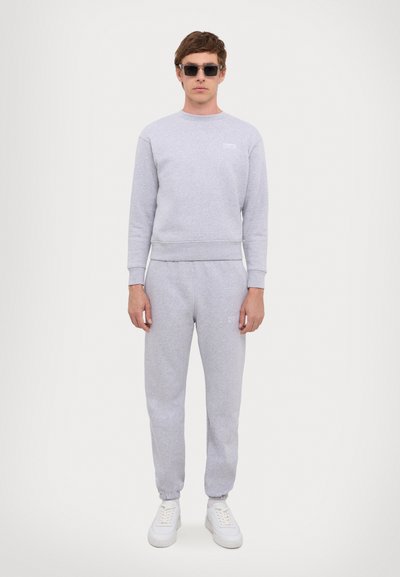 Conjunto de sudadera y pantalones de chándal grises con puños acanalados, letras blancas en el pecho, combinado con zapatillas blancas y gafas de sol negras.
