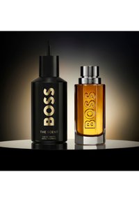 Schwarze Nachfüllflasche und bernsteinfarbene Glasparfümflasche von BOSS The Scent Eau de Toilette auf reflektierender Oberfläche mit dunklem Farbverlauf im Hintergrund.