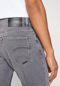 Šedé denimové džíny s texturovaným povrchem, které mají zadní kapsy, výrazné švy a černou koženou značkovou nášivku na pasu.